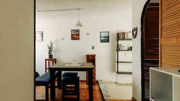 A room with a wooden dining table, two chairs, a small stool, a bookshelf, and a potted plant.