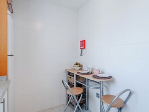 Een compacte kitchenette met een witte betegelde spatwand, een rode brandveiligheidsapparaat, een klein tafeltje met een fruitschaal, twee stoelen en een metalen rek met manden.