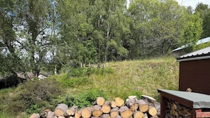 Uma pilha de lenha numa área gramada, com um muro de pedra e uma construção de madeira ao fundo.