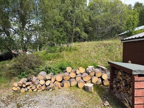 Ein Holzstoß in einer Wiese mit einer Steinmauer und einem Holzbau im Hintergrund.