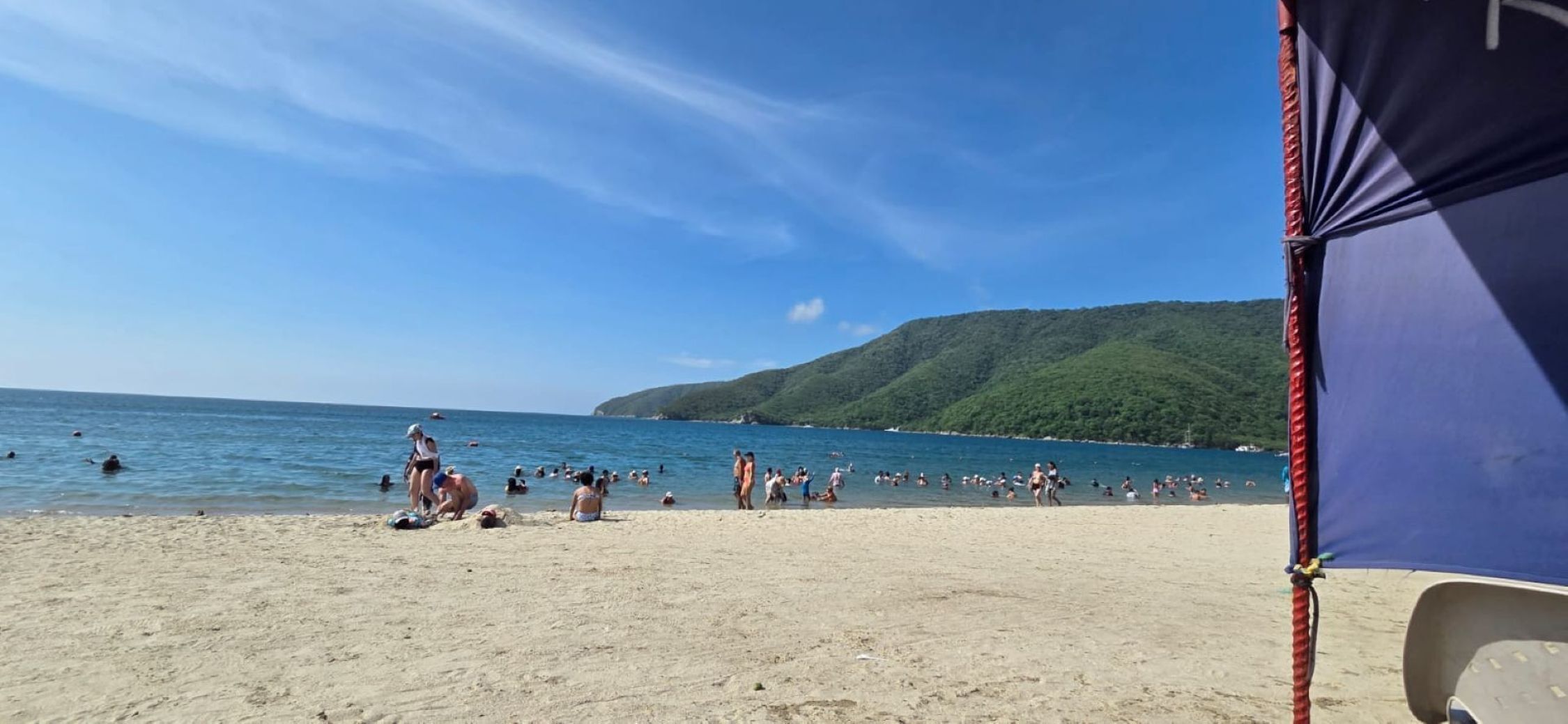 Ein Strand mit badenden und sonnenden Menschen, einem klaren Himmel und einer hügeligen Landschaft im Hintergrund.