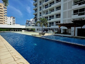 Una piscina con superficie de agua azul cristalina, rodeada por un edificio moderno con múltiples balcones y una zona de estar cubierta.