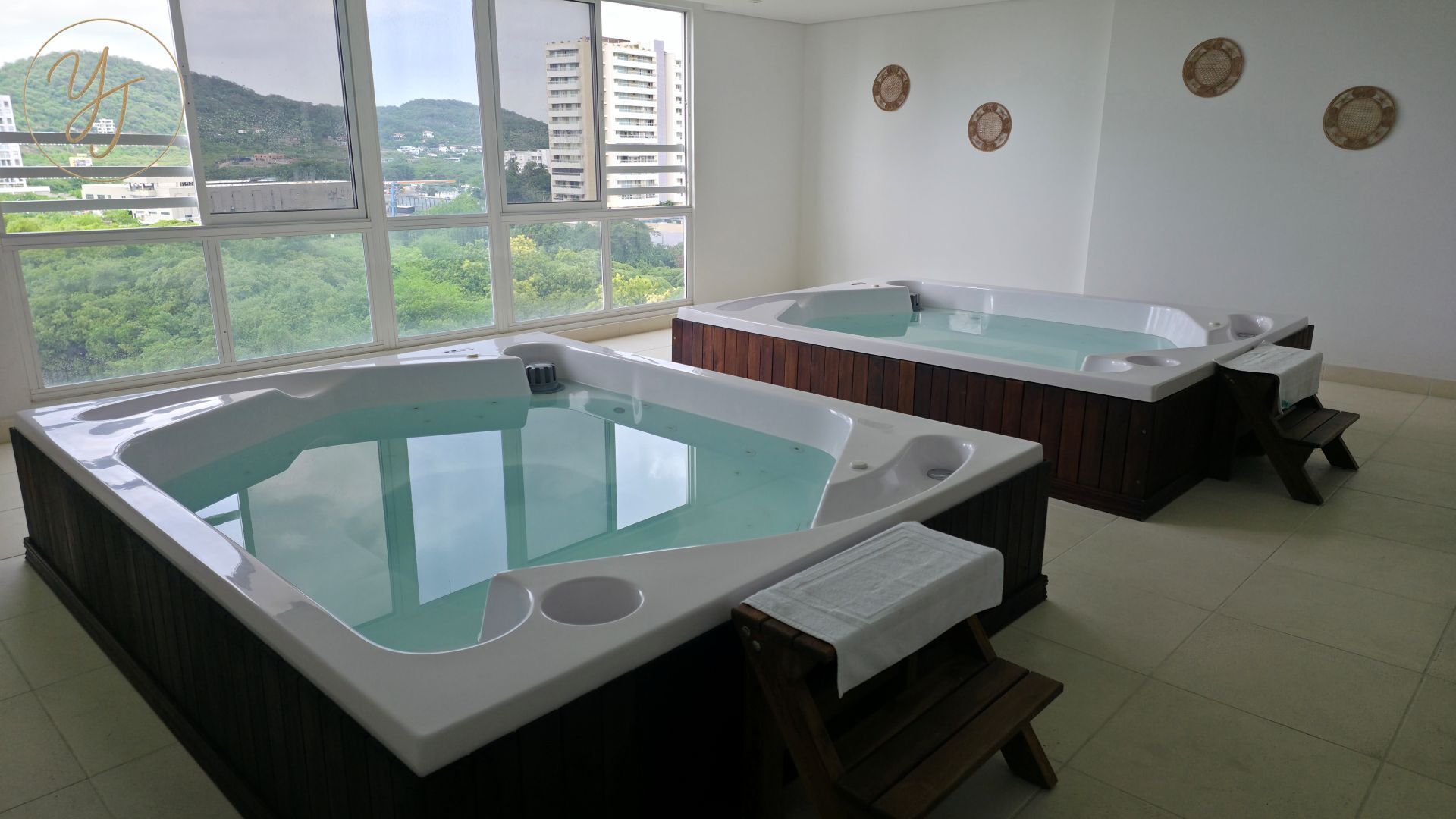 Zwei Whirlpools in einem Raum mit großen Fenstern, Blick auf Grünflächen und Gebäude, sowie eine weiße Wand mit dekorativen Tellern.