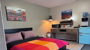 Un dormitorio con cama, escritorio, dispensador de agua y obras de arte en las paredes.