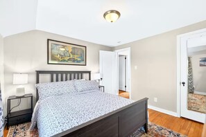 A bedroom with a bed, bedside tables, a painting, and a door leading to another room. - Newly renovated 2-bedrm house in quiet neighborhood, close to everything! (Pittsburgh)
