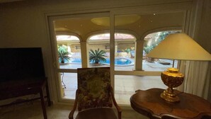 A room with a television, a chair, a lamp, and a view of an outdoor courtyard with a fountain. - Cozy Villa with private pool (Al Shorouk City)