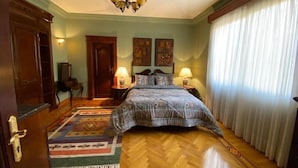 A bedroom with a wooden floor, a bed with a patterned bedspread, two bedside lamps, a chandelier, and a large window with curtains. - Cozy Villa with private pool (Al Shorouk City)