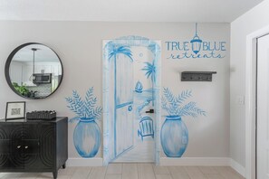 A hallway with a mirrored wall, a black console table with a framed picture, and a door with a blue doorbell and a sign reading 'TRUE BLUE retreats'. - Heated Pool | Hot Tub | Enjoy Buccaneers & Rays (Tampa)