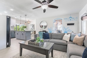A modern living room with a grey sofa, a coffee table, and a ceiling fan. There is a kitchen area with grey cabinets and a dining table. - Heated Pool | Hot Tub | Enjoy Buccaneers & Rays (Tampa)