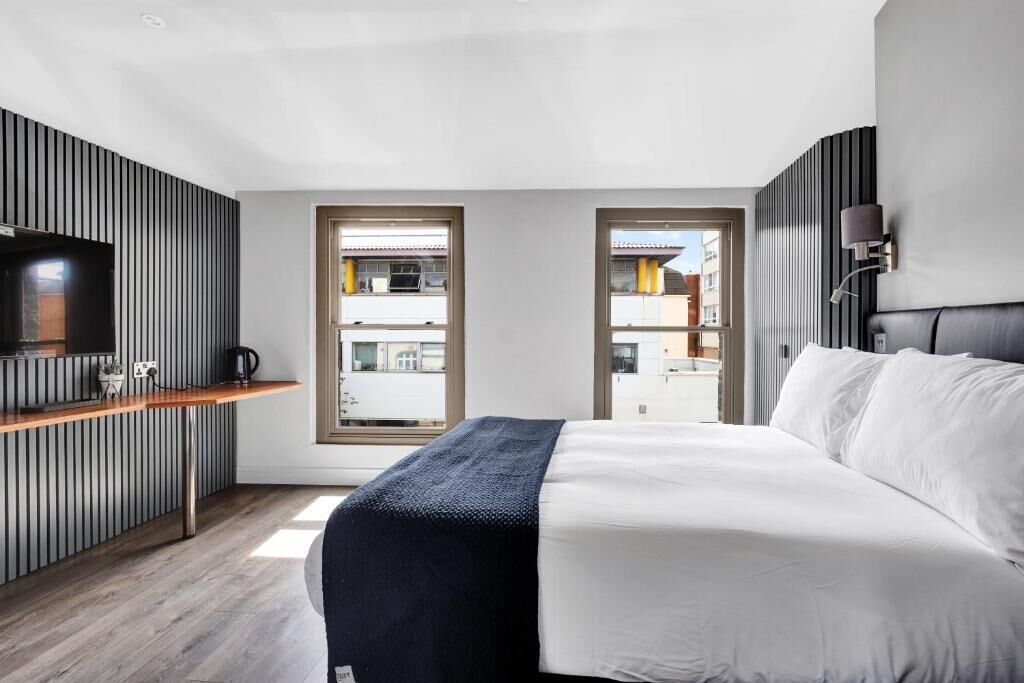 Une chambre d’hôtel moderne équipée d’un lit, d’une télévision, d’une petite kitchenette et de grandes fenêtres.