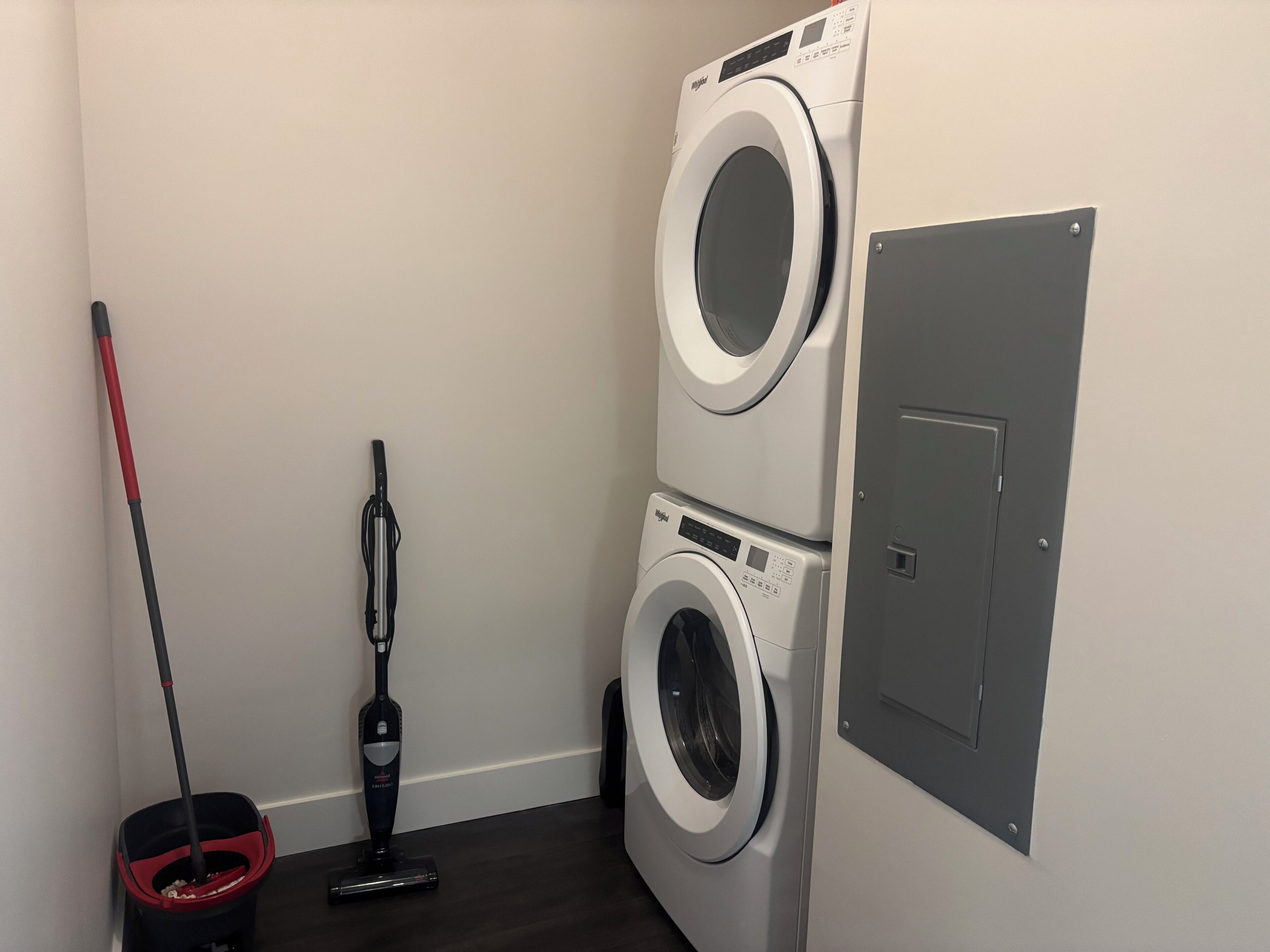 A laundry room with two stacked washing machines, a vacuum cleaner, and a mop.