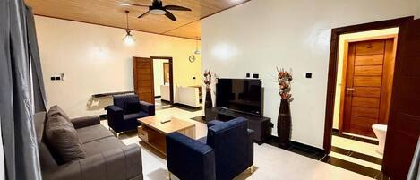 A living room with a sofa, a coffee table, and a TV. There is a ceiling fan and a door leading to another room.