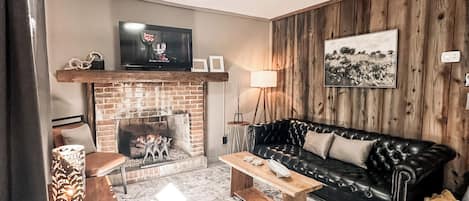A living room with a fireplace, a black leather sofa, a patterned rug, and a television mounted on the wall.