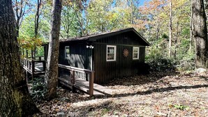Un chalet rustique doté d’une terrasse en bois et d’un petit porche, entouré d’arbres et d’un sol recouvert de feuilles.