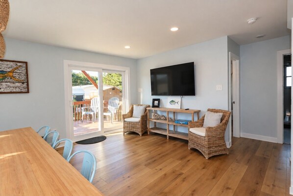 A modern living room with a wooden floor, a flat-screen TV mounted on the wall, wicker furniture, and a sliding glass door leading to an outdoor area.