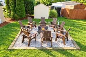 A fire pit surrounded by six Adirondack chairs in a garden area.