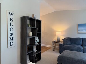 A living room with a dark wooden bookshelf, a white wall with a 'WELCOME' sign, a lamp on a side table, and a blue sofa.