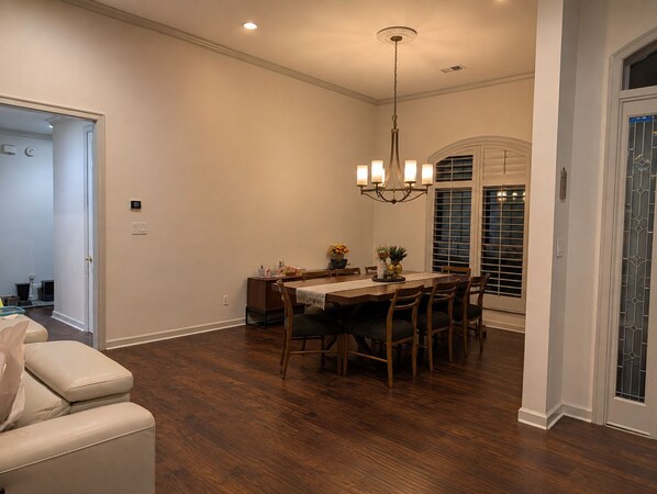 A dining area with a wooden table and chairs, a chandelier, and a window with blinds.