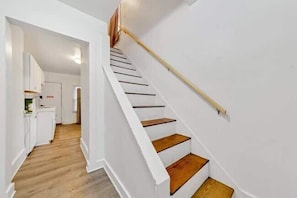 Eine Treppe mit Holzbodenstufen und weißen Wänden, die zu einem Raum führt, im dessen Hintergrund eine Küche erkennbar ist.