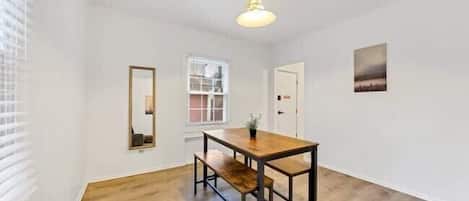 Una habitación con una mesa de madera, bancos, una lámpara colgante, una ventana con persianas y un cuadro en la pared.