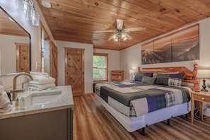Ein Schlafzimmer mit Holzbalkendecke, einem Bett mit Quiltdecke, einem Nachttisch, einer Kommode, einem Spiegel und einem Fenster.