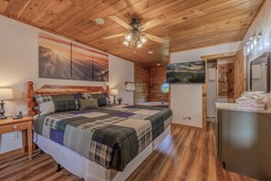 Une chambre avec un plafond en bois, un lit recouvert d’une courtepointe, une table de chevet, une lampe fixée au mur et un tableau représentant un paysage.