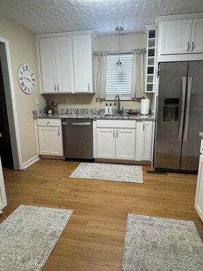 A kitchen with white cabinets, a stainless steel refrigerator, a clock, and a window with blinds.