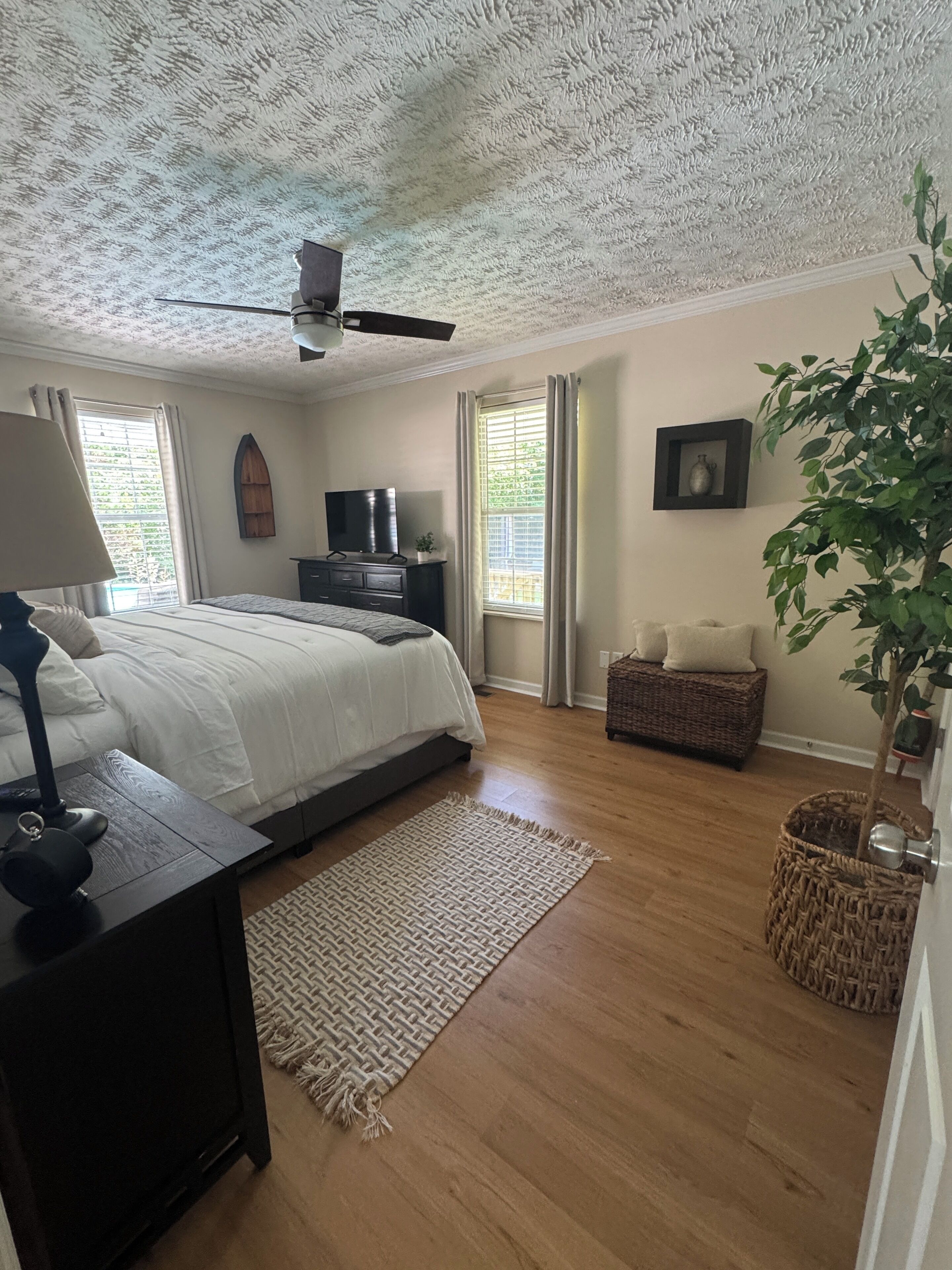 Een slaapkamer met een bed, een dressoir, een televisie, een plafondventilator, een raam met gordijnen en een plant.