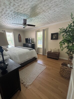 Ein Schlafzimmer mit einem Bett, einem Schminktisch, einem Fernseher, einem Deckenventilator, einem Fenster mit Vorhängen und einer Pflanze.