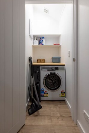 A laundry room with a front-loading washing machine, a vacuum cleaner, and a shelf with cleaning supplies. - Wildberry Cottage - Modern Country Escape (Hamurana)