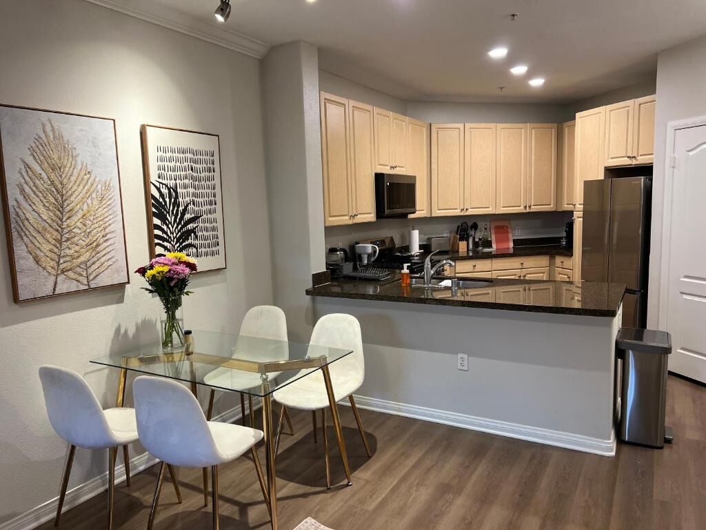 A modern kitchen with wooden cabinets, a stainless steel refrigerator, and a glass dining table with white chairs.