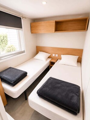 Ein kleines Zimmer mit zwei Einzelbetten, einem Fenster und einem Holzregal darüber.