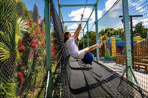 Eine Person auf einer Seilbrücke mit Netzumrandung, umgeben von Grünpflanzen und einer Spielgerüst.
