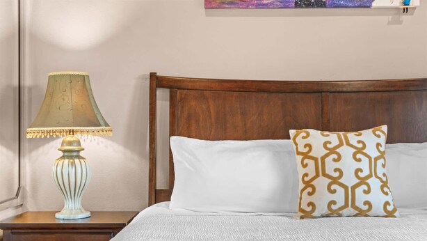 A neatly made bed with white bedding and a decorative pillow, a wooden nightstand with a lamp, and a wooden headboard. - Yakov's Pointe - Pool Access (Hollister)