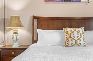 Una cama bien hecha con ropa blanca y una almohada decorativa, una mesita de noche de madera con una lámpara, y un cabecero de madera.