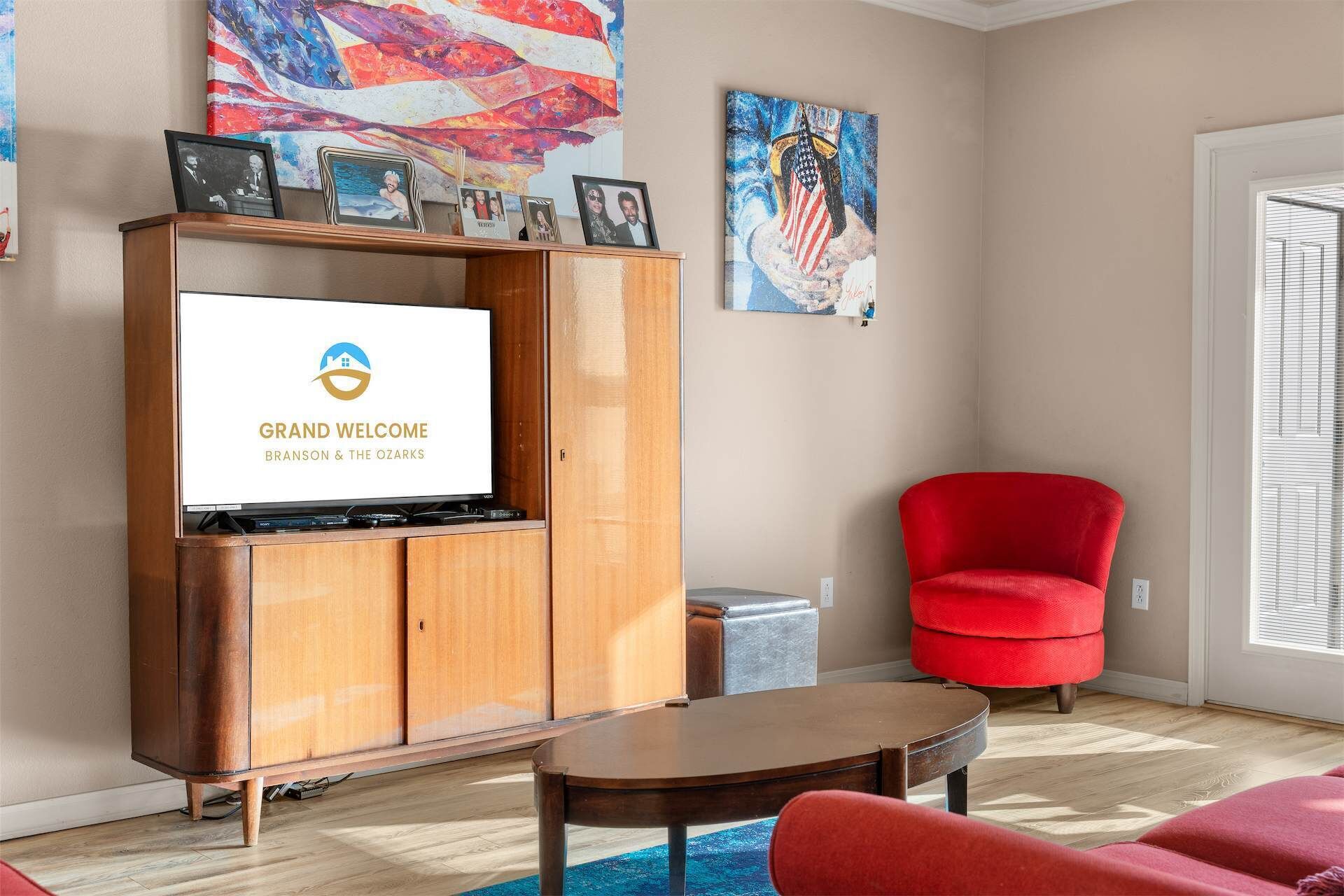 A living room with a television displaying 'GRAND WELCOME BRISON & THE OZARKS', a red armchair, a wooden cabinet, and artwork on the walls.