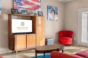 A living room with a television displaying 'GRAND WELCOME BRISON & THE OZARKS', a red armchair, a wooden cabinet, and artwork on the walls. - Yakov's Pointe - Pool Access (Hollister)
