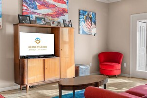 Una sala de estar con una televisión mostrando 'GRAND WELCOME BRISON & THE OZARKS', un sillón rojo, un mueble de madera y obras de arte en las paredes.