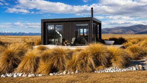 A modern cabin with a large glass door, outdoor seating, and a view of mountains.