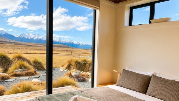 A bedroom with a large window offering a mountain view, a bed with a green blanket, and a white towel.