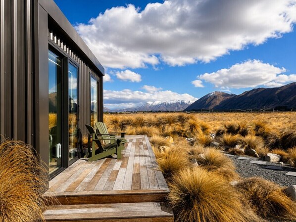 A modern building with a wooden deck and chairs, surrounded by dry grass and mountains in the background.
