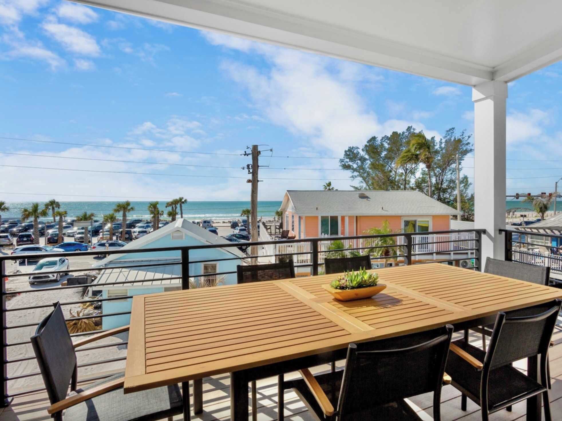 A wooden table with chairs on a balcony overlooking a beach and parking area.