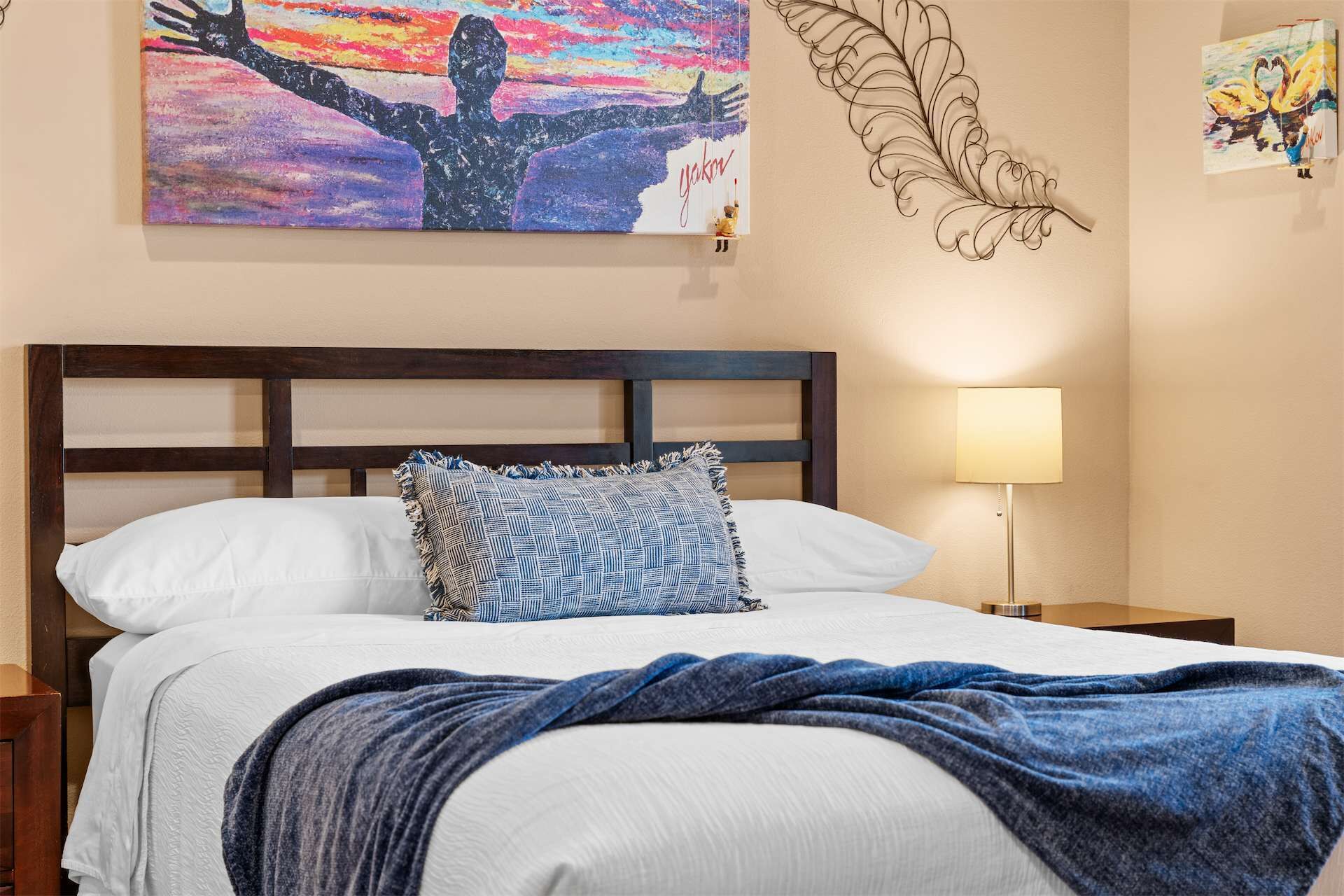 Ein ordentlich bezogenes Bett mit einer blauen Decke, ein Kissen mit blau-weißem Muster und ein Gemälde einer Person mit erhobenen Armen an der Wand.