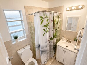 Ein Badezimmer mit Toilette, Waschbecken, Spiegel und einer Duschkabine mit Pflanzenmuster, sowie einem Fenster.