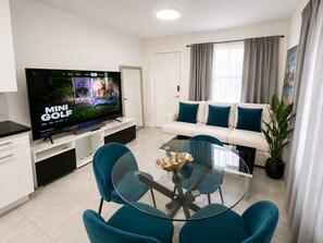 Ein modernes Wohnzimmer mit Flachbildfernseher, einer Couch mit türkisfarbenen Kissen, einem Glastisch und einem Essbereich mit türkisfarbenen Stühlen.