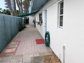 Ein schmaler Pfad führt zu einem Gebäude mit einer blauen Tür, einer weißen Wand und einem daran gelegten, grünen Gartenschlauch.