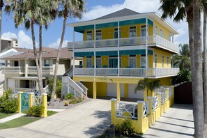 A multi-story yellow building with balconies, a covered entrance, and a parking area with a palm tree. - Canary by the Sea | Sunny Beachside Escape • Private Pool • 2 Min Walk to Beach (South Padre Island)