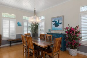 A dining area with a wooden table, chairs, a chandelier, a painting of a sea turtle, and a potted plant with pink flowers. - Canary by the Sea | Sunny Beachside Escape • Private Pool • 2 Min Walk to Beach (South Padre Island)