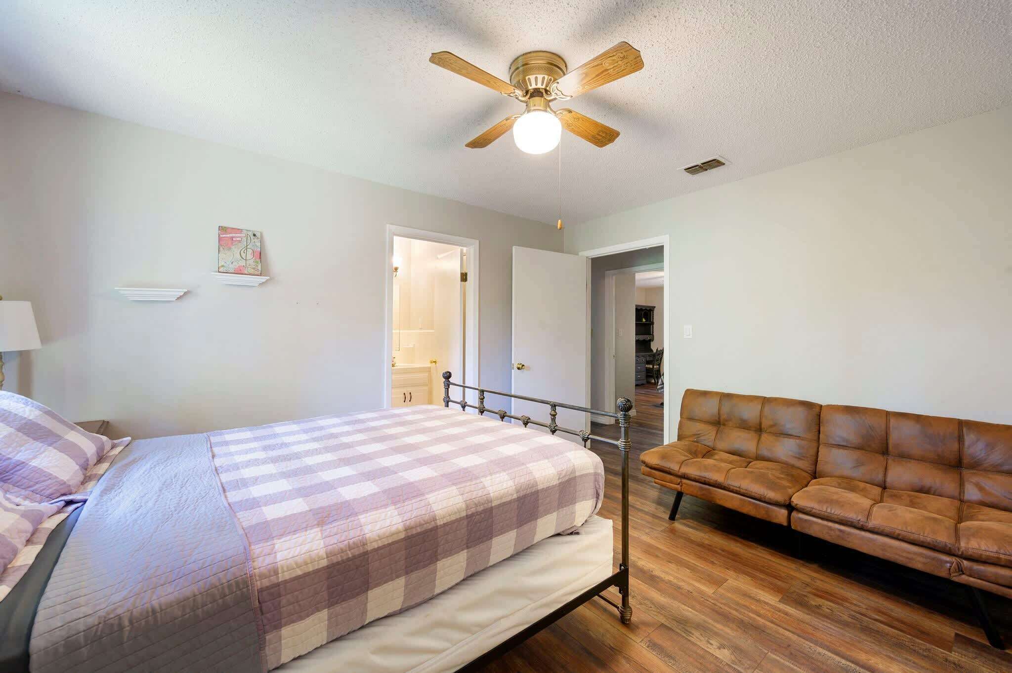 Un dormitorio con una cama, un sofá de cuero marrón, un ventilador de techo y una puerta que da a otra habitación.
