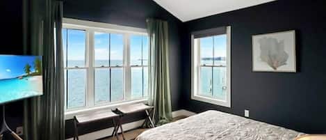 A bedroom with a large bed, a desk, and a view of the sea through a window.